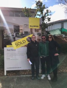 Cate Bakos and two other persons standing in front of a 'Sold' sign for a Ray White property auction, celebrating a successful home purchase. The group poses happily in front of a modern home, with clear skies in the background.